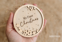 Wooden ornament with 'My First Christmas' text held in a hand against a beige background. Baby shower gift.
