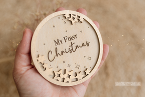 Wooden ornament with 'My First Christmas' text held in a hand against a beige background. Baby shower gift.