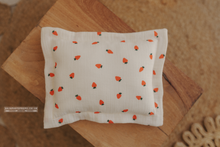 A rectangular newborn posing pillow with a cream color and decorated with a pattern of small orange carrots and white flowers, displayed on a wooden surface.