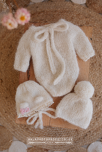A creamy off white baby newborn knitted romper with a beanie and a bear bonnet, displayed on a wooden surface with a floral prop in the background.