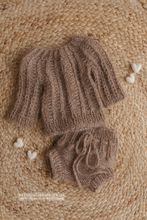 Brown knitted baby hat and booties on a textured beige surface with small white hearts.