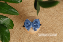 A blue velvet bow tieback for newborns or sitter girls, displayed on a wooden round prop stand, with a green plant in the background.