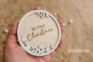 Wooden ornament with 'My First Christmas' text held in a hand against a blurred background. Engraved on a disc.