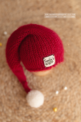 Red knitted Santa hat with a pom-pom on a textured beige background, santa patch, branded 'AAJAPHOTOPROPS.CO.UK'.