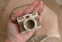 Hand holding a small wooden camera-shaped object with 'Say Cheese!' text.