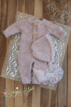 A newborn baby knitted romper and bonnet set in pastel blush pink, displayed on a wooden surface.